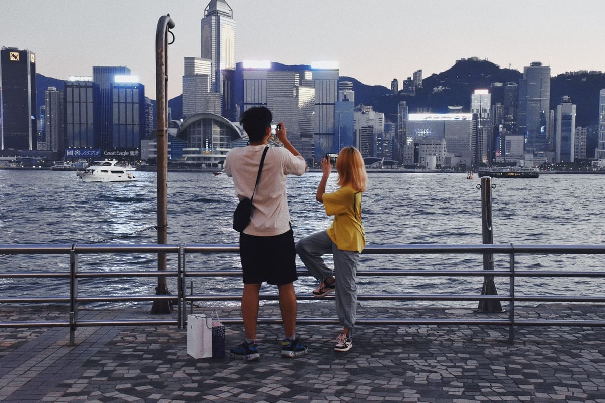 A man and a woman taking photos of Victoria Harbour at dusk with smartphones.